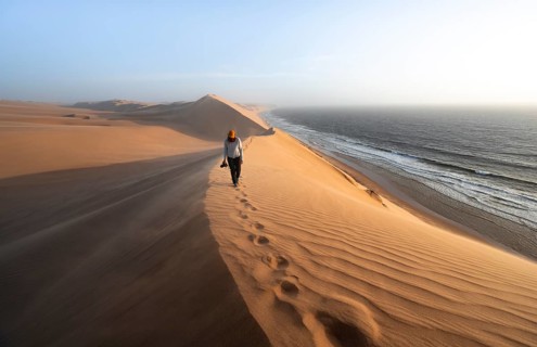Young woman with camera, sand dunes with wave pattern and sea in evening light, wind blowing sand