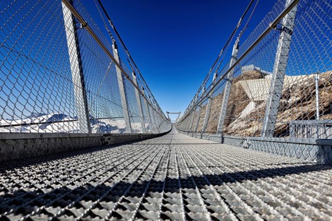 View of a metal suspension bridge against a clear blue sky in a mountainous landscape, The