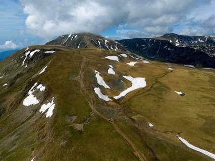 A trail runs through snow-covered mountain peaks under a cloudy sky, aerial view, Transalpina High