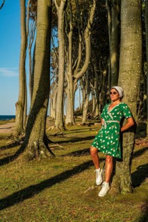 A woman (54) in a green tennis dress and white trainers is leaning casually against a tree, gazing