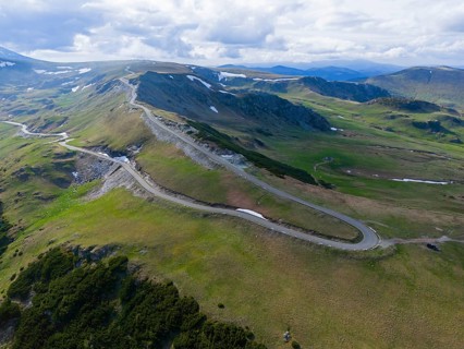 Narrow road snakes through vast green mountains under a slightly cloudy sky, aerial view, near