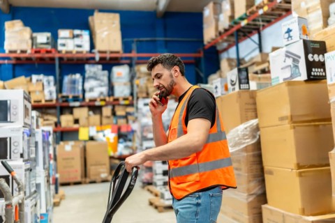 Warehouse worker wearing a safety vest talking on a mobile phone while pulling a pallet jack,