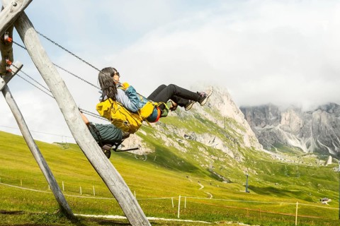 Excited woman with backpack swinging on wooden playground swing lush green hills steep mountain