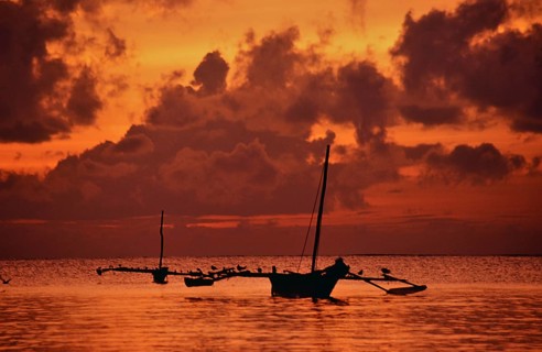 Two traditional wooden boats lie in the calm water off Diani Beach during an intense sunrise. The
