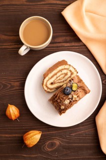 Sweet loaf with cream and caramel, cup of coffee, on brown wooden background with orange textile,
