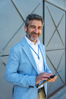 Mature businessman wearing a light blue suit and id badge, holding a digital tablet and smiling,