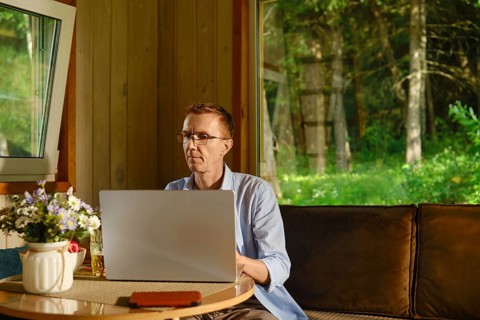 Middle-aged man sits at table using laptop inside wooden cabin and focused on online learning.