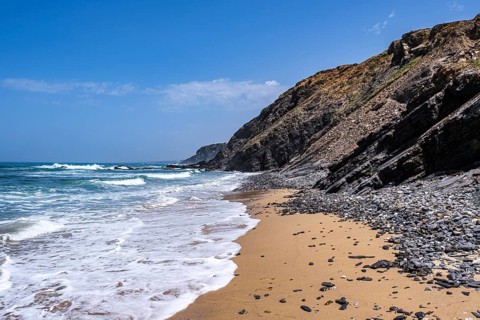 Foam crowns break on a golden sandy beach, with rocky cliffs in the background and clear sky,