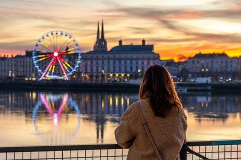 Woman standing on a bridge, overlooking the nive river with the illuminated ferris wheel and city