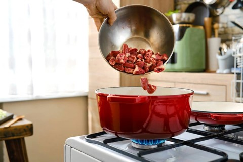 A person pours raw chicken hearts from a bowl into a red cooking pot on a stove in a kitchen.