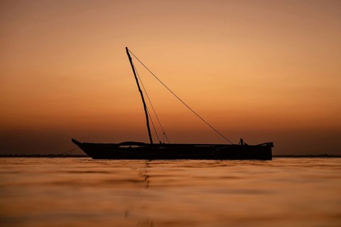A traditional wooden dhow sailing boat is silhouetted against a calm sea and a deep orange sky
