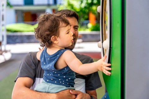 Father holding his young son, with curly hair and a patterned top, engaging with an interactive