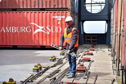 Worker wearing helmet and safety vest holding a large wrench between containers in port, worker