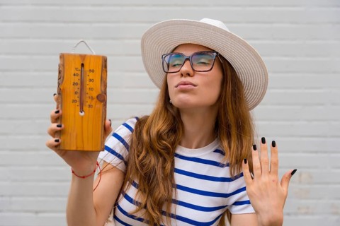 Woman in a hat and sunglasses holding a natural wood thermometer showing high temperature,