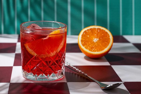A glass fills with a red drink sits on a checkered table. An orange is cut in half next to the