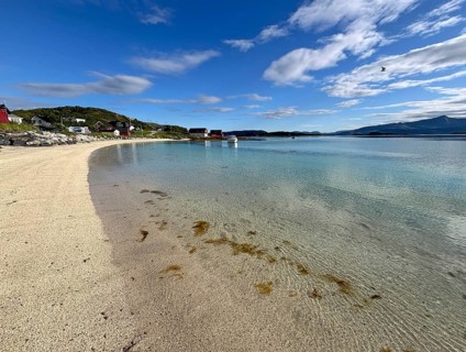 A quiet sandy beach with clear water and a few houses on the shore under slightly cloudy sky,