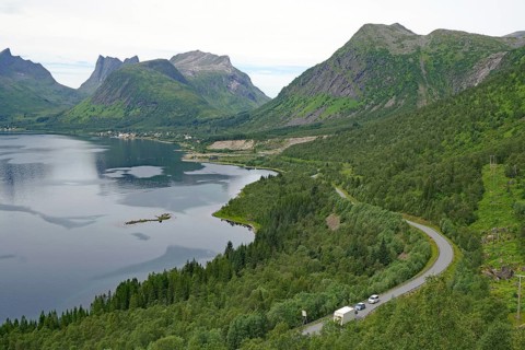 A winding road leads through a green landscape with mountains in the background, a few cars on the