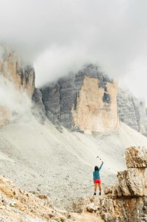 Female hiker raising trekking poles beneath misty Dolomites cliffs celebrating achievement