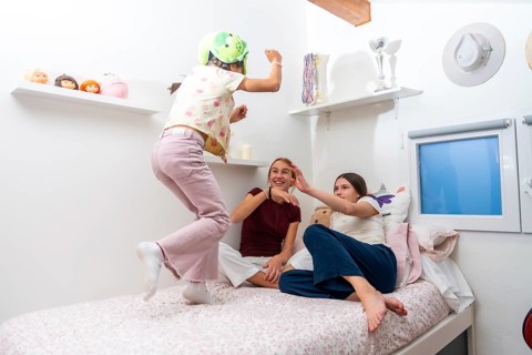 Young sisters happily playing on a bed in a bedroom, one girl wearing a helmet and jumping while