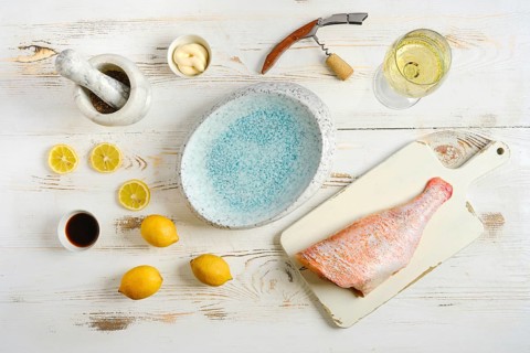 Red snapper lies on cutting board next to lemons, soy sauce, and mortar. Nearby, glass of white