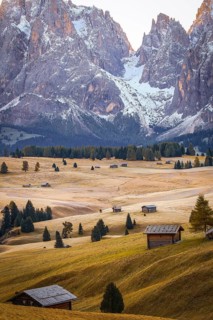Snow-covered mountains frame a vast, quiet meadow landscape with huts, views over the autumnal