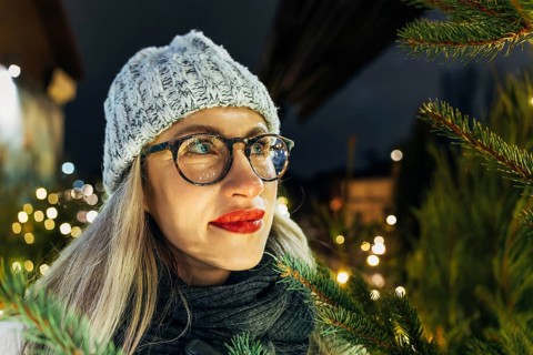 Young woman in glasses among fir branches and lights at night. A woman wearing glasses, a gray knit