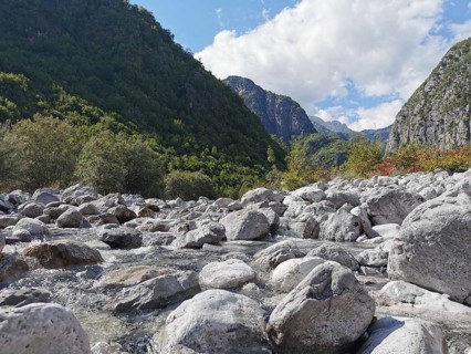 View over a rocky riverbed in a green valley with mountains and cloudy sky, hiking in the Albanian