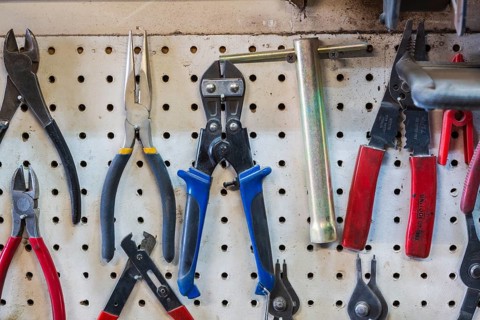 Close-up of various pliers, wire stripper hanging on white pegboard wall in home garage workshop,