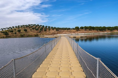 A floating jetty leads across calm water surrounded by green hills under a blue sky, Moura, Beja,