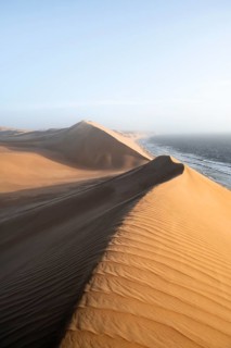 Sand dunes with wave patterns and sea in evening light, wind blowing sand across the dunes,