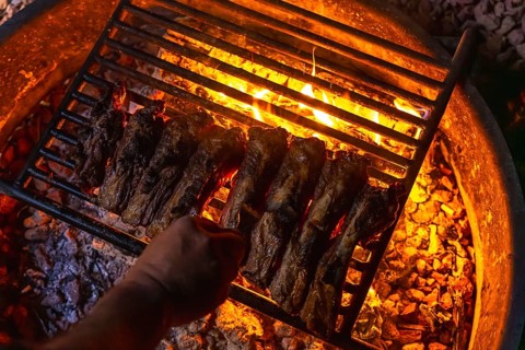 Hand grilling marinated meat ribs over hot flaming coals in a round fire pit, capturing the essence