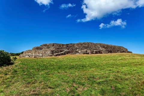 An old stone hill surrounded by a meadow under a clear blue sky with few clouds, The Cairn de