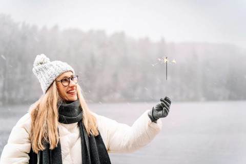 Woman holding a lit sparkler by a snowy lake on a festive winter day