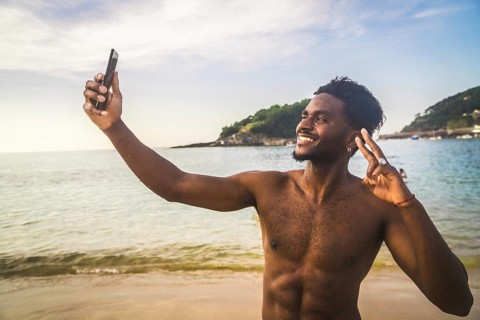 Smiling young man making a peace sign while taking a selfie with a smartphone on a beach,