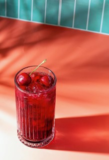 A glass filled with a cherry drink sits on a smooth surface. The drink contains ice and cherries on