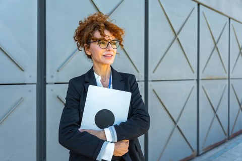 Successful businesswoman in a black suit and glasses holds a laptop, looking away confidently