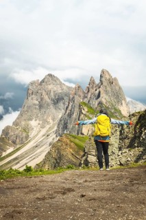Back view middle aged gray hair woman hiker arms outstretched with yellow backpack overlooking lush