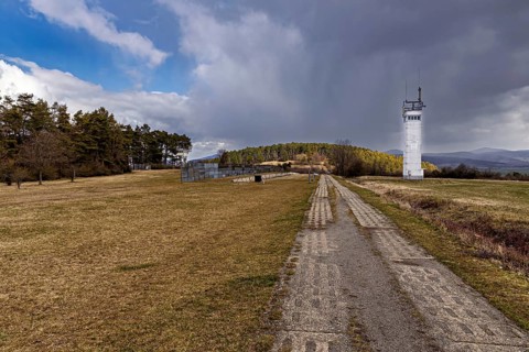 A long way leads to a white watchtower in a meadow with hills in the background, the Point Alpha