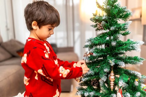 Happy child in red pajamas carefully hangs golden ornaments on a small christmas tree at home,