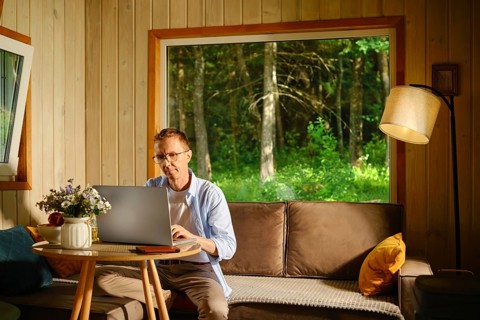 Middle-aged man sits at small round table with laptop in living room in wooden cabin. Sunlight
