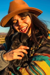 Woman in a brown hat and colorful poncho laughing and hugging a small black puppy at sunrise in