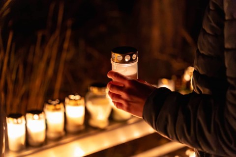 A person holds a lit memorial candle in a glass container outdoors at night, with rows of glowing