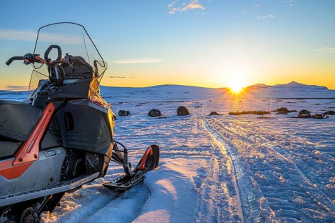 A snowmobile next to tracks in the snow at sunset with mountain views in the background on Sweden's