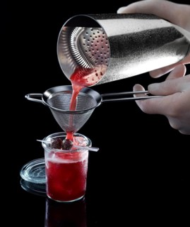 Red cocktail is poured through a sieve, against a black background, 2 berries as garnish, studio