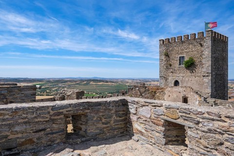 Castle walls with tower and flag offer a wide view of the surrounding countryside under blue skies,