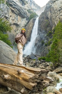 Woman stands on a fallen log, surveying lower yosemite falls and surrounding mountains, forests,