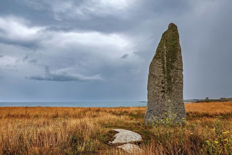 A menhir rises in a meadowy coastal landscape with a view of the sea under dramatic clouds, menhir