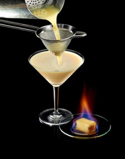 Light brown cocktail is poured through a sieve, against a black background, flambéed confection as