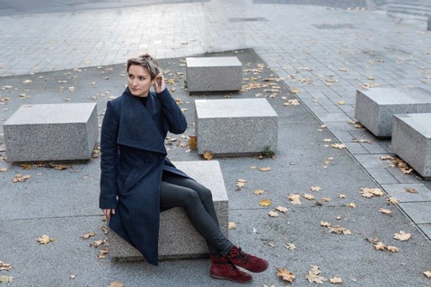 A short-haired attractive blonde in a coat and red shoes sits in the town square. A woman in a dark