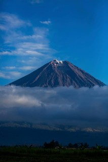 A majestic volcano rises above clouds and a blue sky with light snow cover at the summit, Smoking